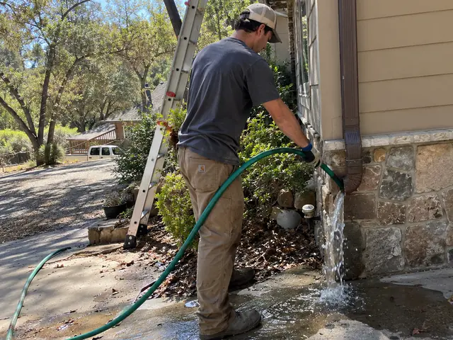 A crew member running a garden hose into a downspout until water pours out the bottom onto the driveway