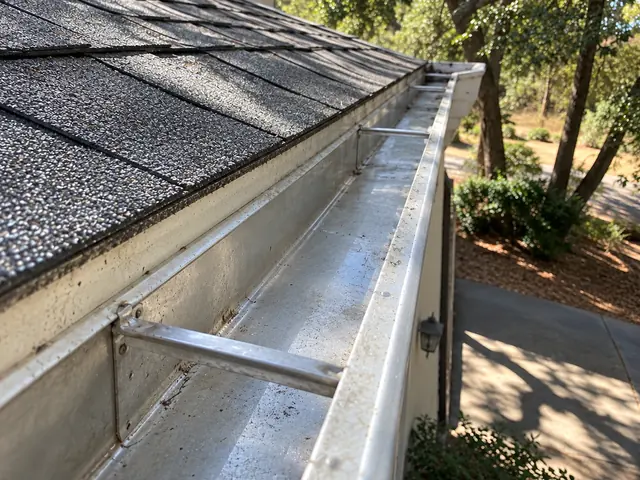 A clean metal gutter seen down its length beside a shingle roof, with oaks and a driveway below