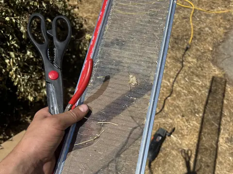 A hand holding a section of metal-framed mesh gutter guard with needles caught on the mesh, beside a pair of scissors